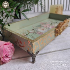 May include: A rectangular sage green tray featuring a floral print on the interior base, gold leaf accents on the exterior sides, and ornate metallic feet. The side of the tray is decorated with a gold floral relief and the text re-design.