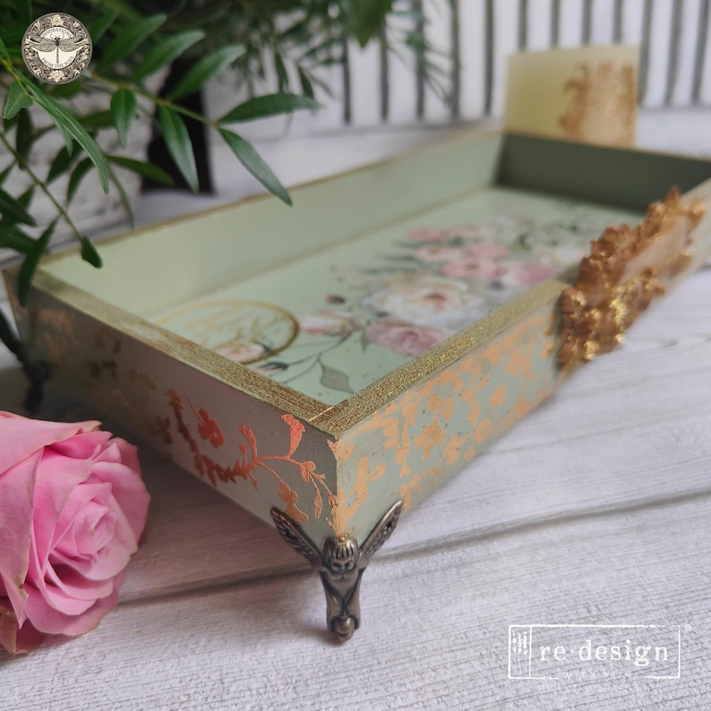 May include: A rectangular sage green tray featuring a floral print on the interior base, gold leaf accents on the exterior sides, and ornate metallic feet. The side of the tray is decorated with a gold floral relief and the text re-design.