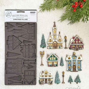 May include: A silicone mold for creating Christmas village decorations, featuring various house and tree designs. The finished decorations are painted in festive colors like red, green, and gold. The mold is labeled "DECOR MOULDS CHRISTMAS VILLAGE".