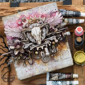 May include: A mixed media artwork on a wooden panel featuring a central ram skull design with wings, gears, and floral elements. The color palette includes white, silver, gold, and touches of pink and brown. The artwork has a steampunk and gothic theme.