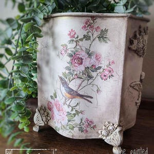 May include: A white ceramic planter with a floral design featuring pink roses, green leaves, and a small brown bird. The planter has a scalloped edge and decorative feet.