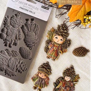 May include: A silicone mold for making pine cone dolls. The mold has five different designs, including a pine cone, a doll with a pine cone hat, and a doll with a pine cone dress. The mold is gray and has the words "Decor Moulds" and "Pinecone Dolls" printed on it.