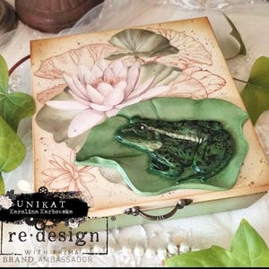 May include: A rectangular wooden box featuring a decorative top with a printed illustration of a pink water lily and green lily pads. A three-dimensional green frog figurine is attached to the lid, resting on a sculpted green lily pad.