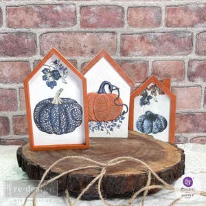 May include: Three wooden framed signs with fall-themed pumpkin illustrations. The signs are displayed on a wooden slice. The signs feature pumpkins with blue and white patterns, a pumpkin with orange and white patterns, and a pumpkin with blue and white patterns.