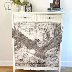 May include: A white cabinet with two drawers and two doors, featuring a distressed eagle graphic. The drawers have a striped design and decorative hardware. The cabinet is adorned with the text "THE ROYAL COURT" and "Valerie DeVincent".