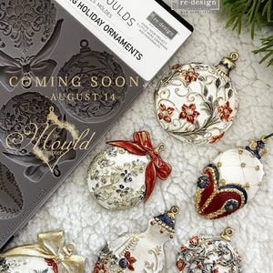 May include: A collection of shimmering holiday ornaments with intricate floral designs in red, gold, and white. The ornaments are displayed on a soft, white surface, alongside a mold package that reads "Coming Soon August 14."