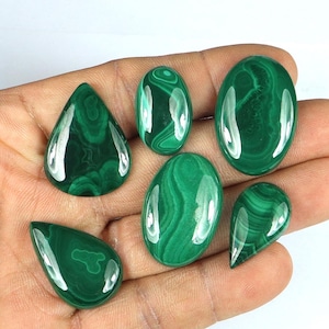 May include: A collection of polished malachite gemstones in various shapes, including oval and teardrop. The gemstones display a rich, deep green color with swirling patterns, reflecting light and showcasing their natural beauty. The gemstones are held in a hand.