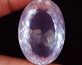 May include: A large, faceted, oval-shaped gemstone with a pale pink hue. The gem is held between fingers and displays a complex pattern of light refraction. The text "71.20 ct" is visible at the top left.