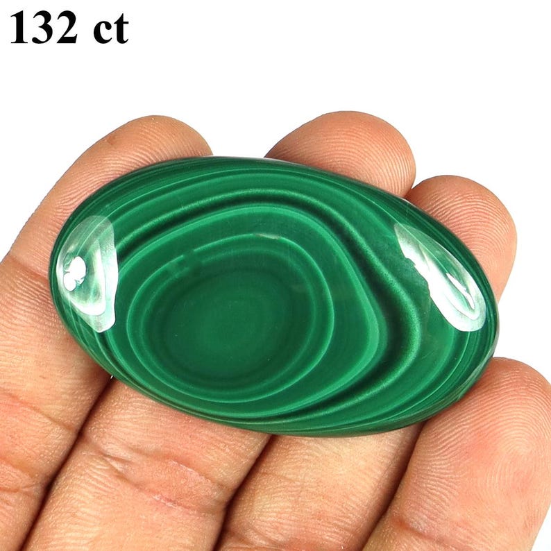 May include: An oval-shaped, polished malachite gemstone with a deep green color and swirling patterns. The stone is held in a hand, and the text "132 ct" is visible in the upper left corner.