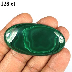 May include: A polished, oval-shaped malachite gemstone with a deep green color and swirling patterns. The stone is held in a hand, and the text "128 ct" is visible in the upper left corner. The gemstone is a natural mineral.