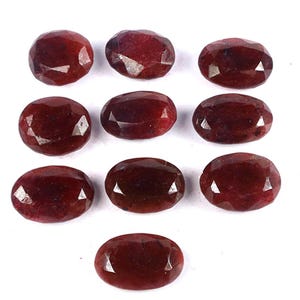 Puede incluir: Diez piedras preciosas rojas de forma ovalada y talladas en facetas, dispuestas en una cuadrícula. Cada piedra presenta una superficie pulida con múltiples facetas triangulares que reflejan la luz, mostrando tonos carmesí profundos.