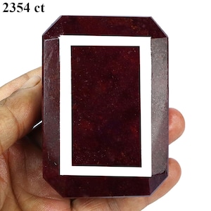 May include: A large, rectangular, dark red gemstone with a white border. The gemstone is held in a hand and has the text "2354 ct" in the upper left corner. The gemstone has a polished surface.