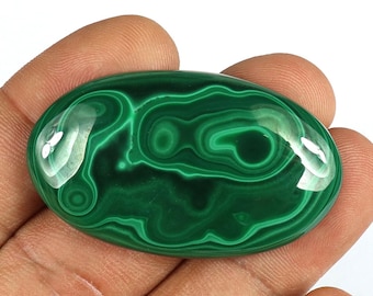 May include: A polished, oval-shaped malachite gemstone with a vibrant green color and swirling patterns. The gemstone is held in a hand and the text "101 ct" is visible in the image.