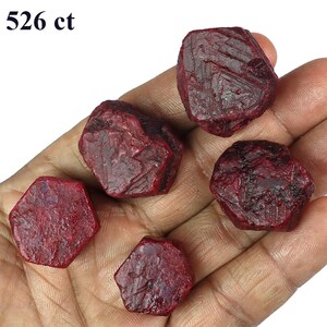 May include: Five rough-cut, deep red ruby gemstones. The stones have a textured surface and irregular shapes. The text "526 ct" is in the upper left corner. These gemstones are suitable for jewelry making or collecting.