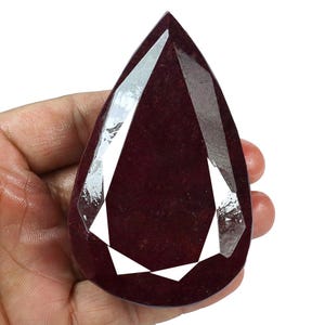 Può includere: Una grande gemma sfaccettata a forma di pera con una tonalità rosso intenso. La gemma ha molteplici sfaccettature che riflettono la luce, creando una brillantezza brillante. La pietra è tenuta in mano su sfondo bianco.