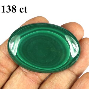 May include: A polished, oval-shaped malachite gemstone, displaying concentric bands of dark and light green. The stone is held in a hand, and the text "138 ct" is visible in the upper left corner. The gemstone has a glossy, reflective surface.