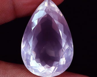 May include: A pear-shaped, faceted gemstone with a pale pink hue. The gem is held between fingers and has the text "46.85 ct" at the top. The cut of the stone creates multiple reflective surfaces.
