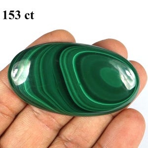 May include: A polished, oval-shaped malachite gemstone, displaying a rich, deep green color with swirling patterns. The stone weighs 153 ct and is held in a hand, showcasing its smooth, glossy surface.