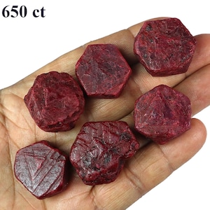 Può includere: Una collezione di sei gemme di rubino grezze, di colore rosso intenso. Le gemme sono tenute in una mano, mostrando la loro consistenza naturale e non lucidata. Il testo "650 ct" è visibile in alto a sinistra.