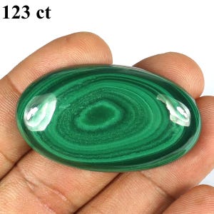 May include: Oval-shaped malachite gemstone with concentric green patterns. The polished stone has a glossy surface and is held in a hand. The text "123 ct" is in the upper left corner.