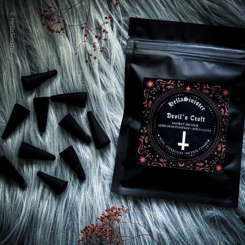 Charcoal incense cone burning on a ceramic incense holder, with a black bag labeled “Devils Croft” pumpkin and clove incense cones by HellaSinister, photographed on a dark gothic background with vintage occult book props.