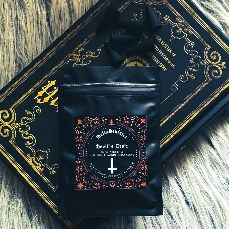Charcoal incense cone burning on a ceramic incense holder, with a black bag labeled “Devils Croft” pumpkin and clove incense cones by HellaSinister, photographed on a dark gothic background with vintage occult book props.