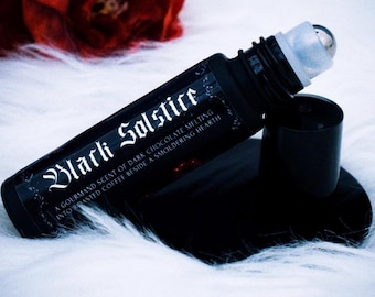 Puede incluir: Botella roll-on negra con el texto "Black Solstice" y una descripción de un aroma gourmand de chocolate negro derritiéndose en café tostado. La botella tiene un aplicador de bola plateada y una tapa negra. El fondo es blanco.