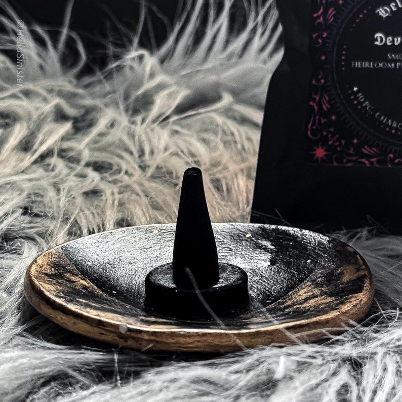 Charcoal incense cone burning on a ceramic incense holder, with a black bag labeled “Devils Croft” pumpkin and clove incense cones by HellaSinister, photographed on a dark gothic background with vintage occult book props.