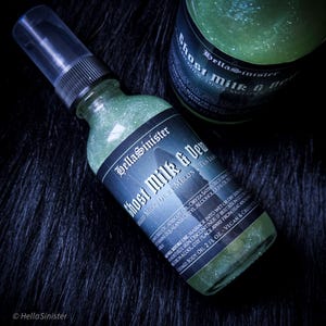 May include: Two green glass bottles of "Ghost Milk & Dew" body oil with a black label. The bottles are on a dark, textured surface. The label includes the brand name "BellaSinister".