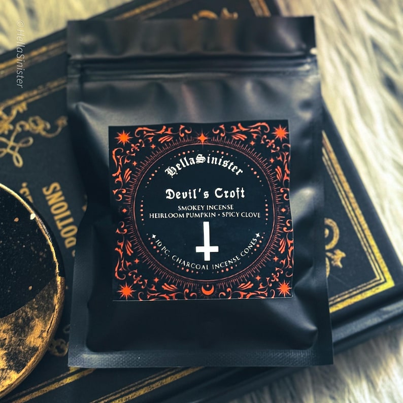 Charcoal incense cone burning on a ceramic incense holder, with a black bag labeled “Devils Croft” pumpkin and clove incense cones by HellaSinister, photographed on a dark gothic background with vintage occult book props.