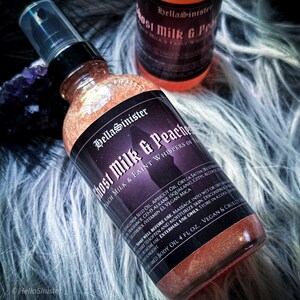 May include: A bottle of shimmering peach-coloured body oil with a black label. The label reads "Ghost Milk & Peaches". The bottle has a black spray nozzle. A second bottle is visible in the background.