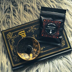 Charcoal incense cone burning on a ceramic incense holder, with a black bag labeled “Devils Croft” pumpkin and clove incense cones by HellaSinister, photographed on a dark gothic background with vintage occult book props.