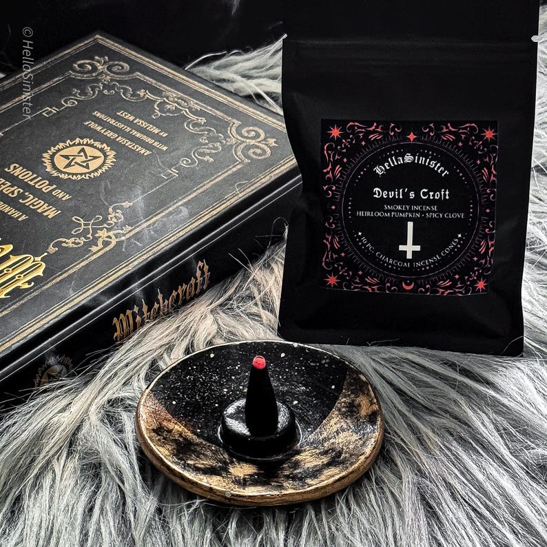 Charcoal incense cone burning on a ceramic incense holder, with a black bag labeled “Devils Croft” pumpkin and clove incense cones by HellaSinister, photographed on a dark gothic background with vintage occult book props.