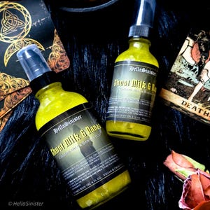 May include: Two bottles of "Ghost Milk & Nanas" perfumed body oil with yellow liquid and black labels. The labels feature the text "HellaSinister" and product information. The bottles are on a dark, textured surface.