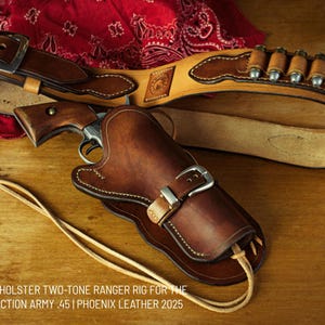 Puede incluir: Un holster de cuero marrón con un diseño de dos tonos, un cinturón de cuero marrón con un bucle de balas y una pistola plateada. El texto "THE VAQUERO HOLSTER TWO-TONE RANGER RIG FOR THE COLT SINGLE ACTION ARMY .45 | PHOENIX LEATHER 2025" está impreso debajo del holster.
