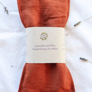 K&ouml;nnte beinhalten: Ein rostfarbenes, gewichtetes Augenpolster aus Leinen mit einem Papierband darum. Auf dem Band steht "Lavender and Flax Weighted Linen Eye Pillow". Getrocknete Lavendelzweige sind um das Kissen auf einer wei&szlig;en Oberfl&auml;che verstreut.