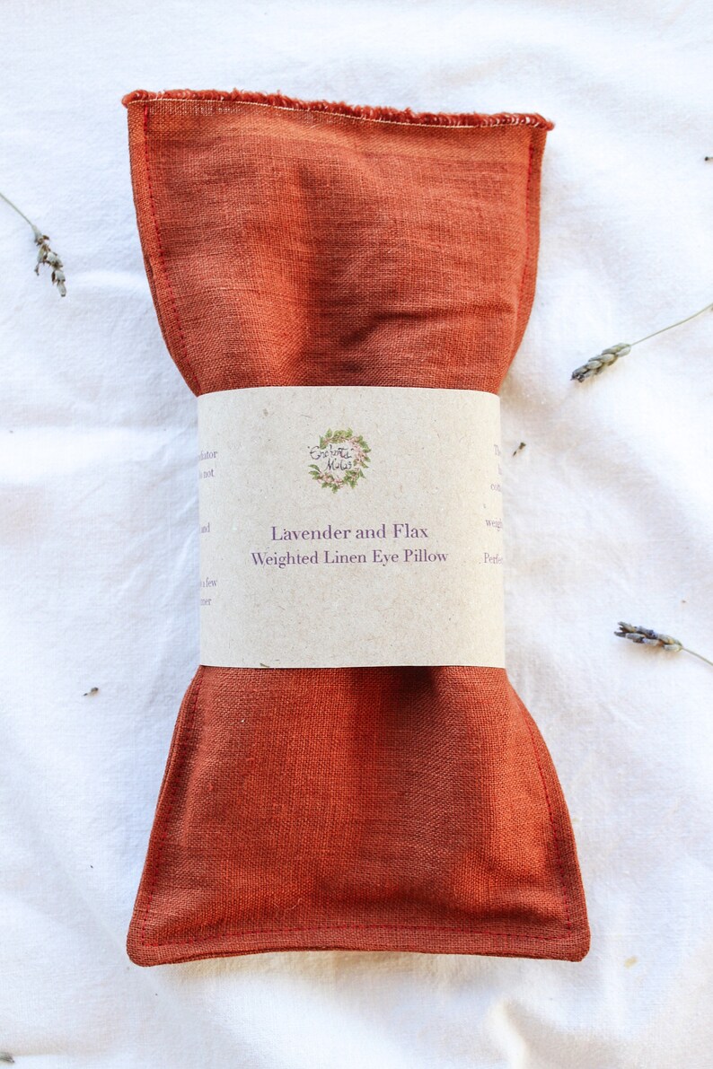 K&ouml;nnte beinhalten: Ein rostfarbenes, gewichtetes Augenpolster aus Leinen mit einem Papierband darum. Auf dem Band steht "Lavender and Flax Weighted Linen Eye Pillow". Getrocknete Lavendelzweige sind um das Kissen auf einer wei&szlig;en Oberfl&auml;che verstreut.