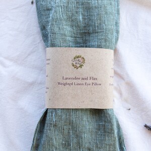 K&ouml;nnte beinhalten: Ein gr&uuml;n-wei&szlig; gestreiftes Leinen-Augenkissen mit einem braunen Papierband. Auf dem Band steht "Lavender and Flax Weighted Linen Eye Pillow". Das Kissen ist gefaltet und gebunden und liegt auf einer wei&szlig;en Oberfl&auml;che.