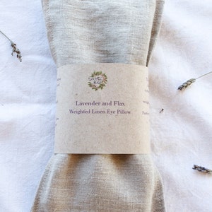 K&ouml;nnte beinhalten: Ein beiges Leinen-Augenkissen mit einem braunen Papierband. Auf dem Band steht "Lavender and Flax Weighted Linen Eye Pillow" mit einem floralen Logo. Getrocknete Lavendelzweige sind um das Kissen auf einer wei&szlig;en Oberfl&auml;che verstreut.