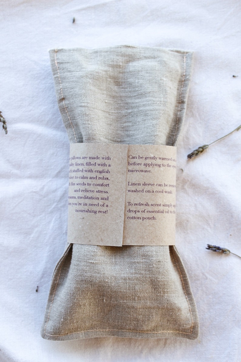 K&ouml;nnte beinhalten: Ein beiges Leinenkissen, das zur Entspannung dient, ist mit einem braunen Papierband mit Text umwickelt. Das Kissen ist mit Leinsamen und englischem Lavendel gef&uuml;llt. Der Text beschreibt die Verwendung und Pflege.
