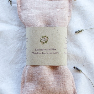 K&ouml;nnte beinhalten: Ein rosa Leinen-Augenkissen mit einem braunen Papierband. Auf dem Band steht "Lavender and Flax Weighted Linen Eye Pillow" und ein Blumenlogo. Getrocknete Lavendelzweige sind um das Kissen auf einem wei&szlig;en Hintergrund verteilt.