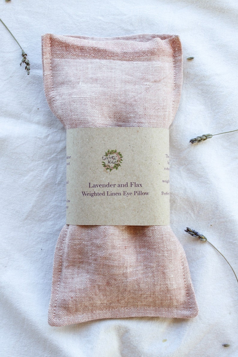 K&ouml;nnte beinhalten: Ein rosa Leinen-Augenkissen mit einem braunen Papierband. Auf dem Band steht "Lavender and Flax Weighted Linen Eye Pillow" und ein Blumenlogo. Getrocknete Lavendelzweige sind um das Kissen auf einem wei&szlig;en Hintergrund verteilt.