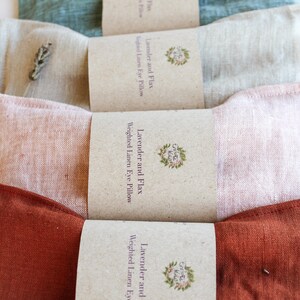 K&ouml;nnte beinhalten: Vier gewichtete Augenpolster aus Leinen in den Farbt&ouml;nen T&uuml;rkis, Beige, Rosa und Rost. Jedes Kissen ist mit einem braunen Papierband mit der Aufschrift "Lavender and Flax Weighted Linen Eye Pillow" und einem floralen Logo versehen. Die Kissen sind in einer Reihe angeordnet.