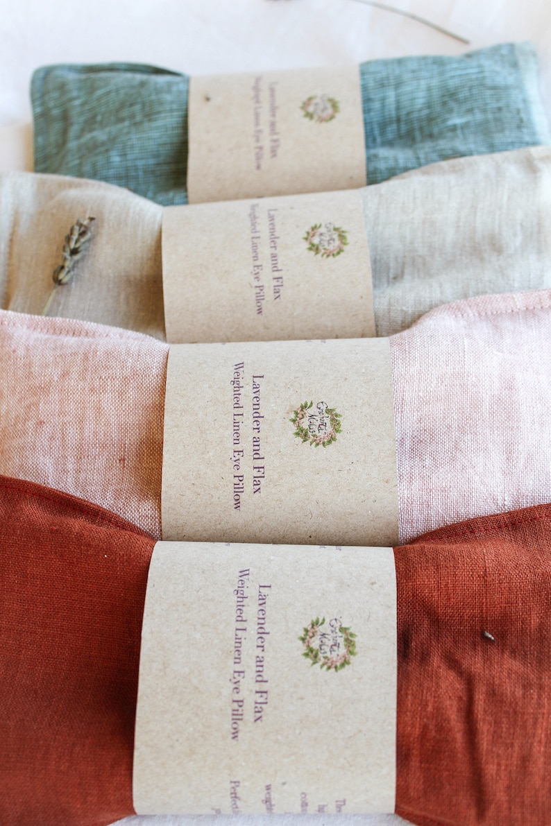 K&ouml;nnte beinhalten: Vier gewichtete Augenpolster aus Leinen in den Farbt&ouml;nen T&uuml;rkis, Beige, Rosa und Rost. Jedes Kissen ist mit einem braunen Papierband mit der Aufschrift "Lavender and Flax Weighted Linen Eye Pillow" und einem floralen Logo versehen. Die Kissen sind in einer Reihe angeordnet.