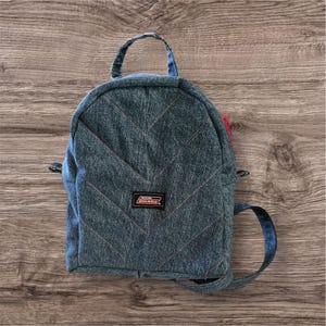 May include: A small, denim backpack with a quilted design and pink stitching. The backpack features a top handle, adjustable straps, and a small Dickies brand patch. The bag is made of blue denim.