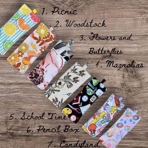 Könnte beinhalten: Sieben Stoff-Stifteetuis mit verschiedenen Mustern und Designs. Die Muster umfassen Fruchtscheiben, Retro-Blumen, Blumenmuster, Schulmaterial und Donuts. Jedes Etui hat eine kleine Schlaufe und den Text: Picnic, Woodstock, Flowers and Butterflies, Magnolias, School Time, Pencil Box und Candyland.