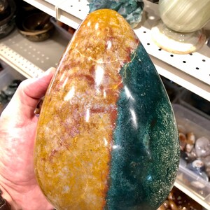 Puede incluir: Una piedra grande, lisa, pulida, en forma de lágrima con un patrón de color verde y amarillo. La piedra está siendo sostenida en una mano.