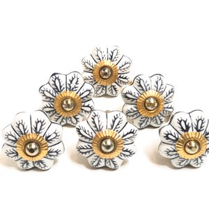 以下が含まれることがあります： 黒い花柄と金色のアクセントが付いた白いセラミック製の引き出しノブが6個。ノブは5枚の花びらを持つ花の形をしています。