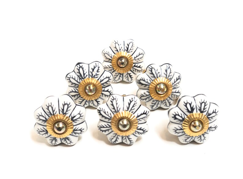 以下が含まれることがあります： 黒い花柄と金色のアクセントが付いた白いセラミック製の引き出しノブが6個。ノブは5枚の花びらを持つ花の形をしています。