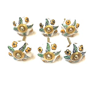 Puede incluir: Seis pomos de cerámica blanca para armarios con diseño floral. Cada pomo presenta una base de metal dorado y una joya central. El patrón floral incluye flores naranjas y hojas verdes.